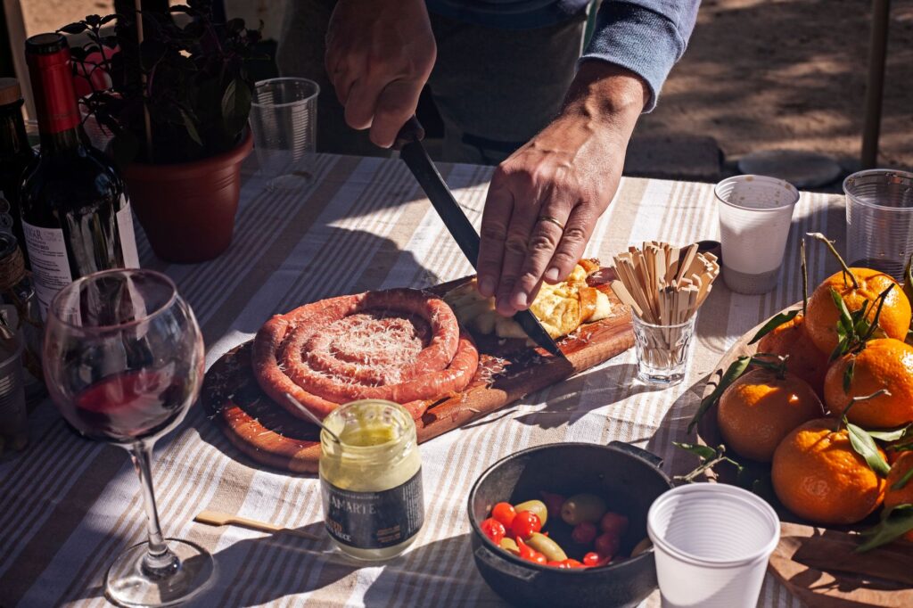Un homme prépare un délicieux repas en plein air avec de la saucisse, des fruits et du vin par une journée ensoleillée.