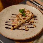Une part de cheesecake crémeux garnie de noix et de menthe, arrosée de sauce au chocolat.