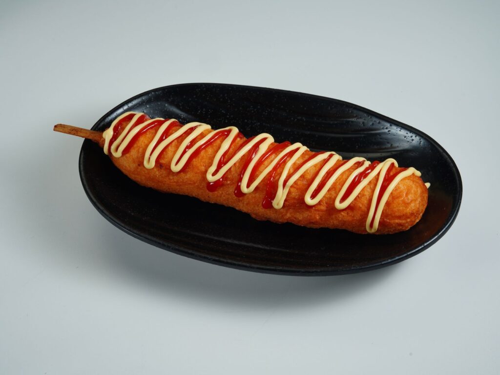 Un corndog parfaitement cuit, garni de ketchup et de mayonnaise sur une assiette noire.