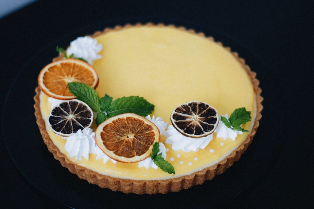 Tarte au citron élégante garnie de tranches d'agrumes séchées et de menthe sur une surface noire.