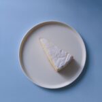 Une seule tranche de gâteau au citron saupoudrée de sucre glace sur une assiette blanche à fond bleu.