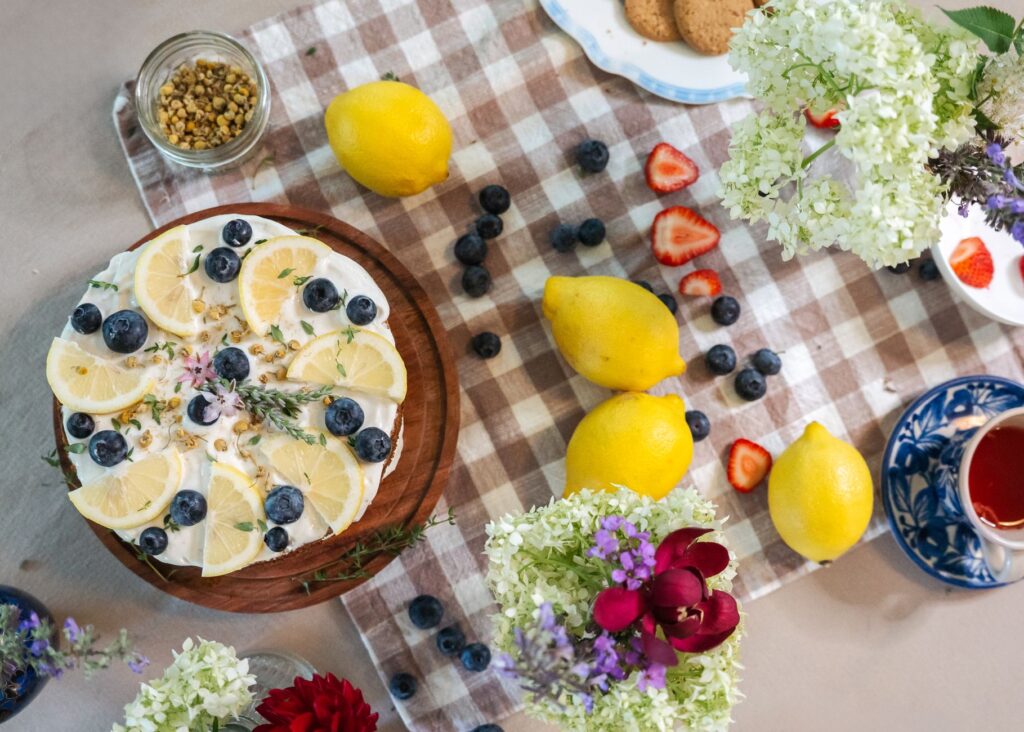 Un délicieux gâteau au citron et aux myrtilles, entouré de fruits frais et de fleurs sur une table.