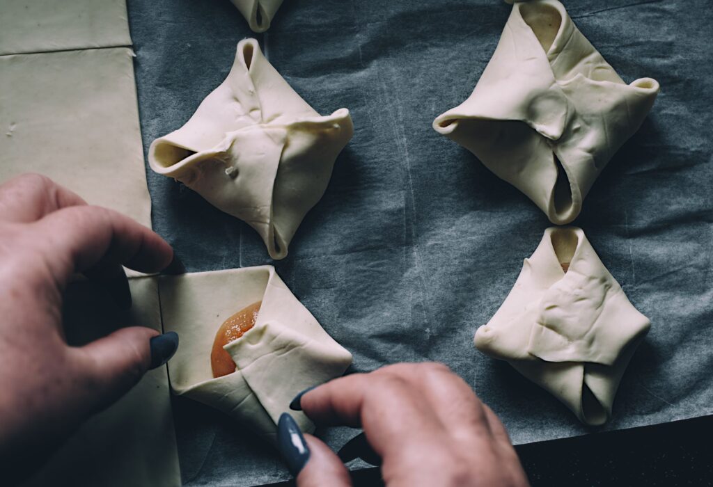 Gros plan sur des mains pliant des feuilletés sur du papier sulfurisé dans une cuisine.