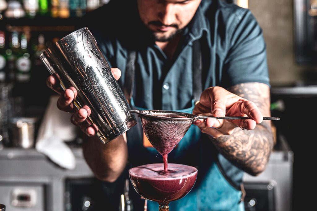 Un barman expérimenté prépare un cocktail à l'aide d'une passoire et d'un shaker dans un bar à l'ambiance animée.