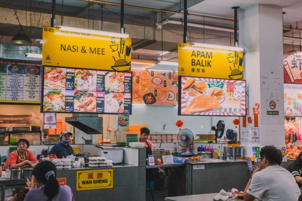 Scène intérieure animée d'une aire de restauration asiatique avec divers stands et des clients locaux savourant leurs repas.