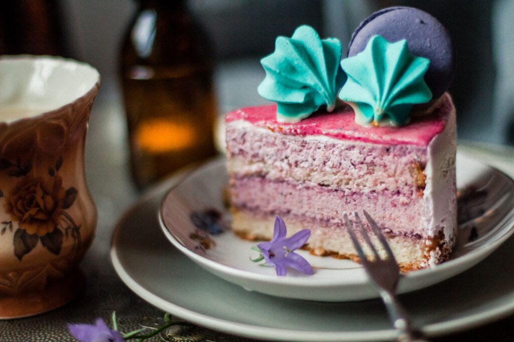 Une tranche de gâteau coloré en couches décorée d'un macaron et de tourbillons de crème vibrants sur une assiette florale classique.