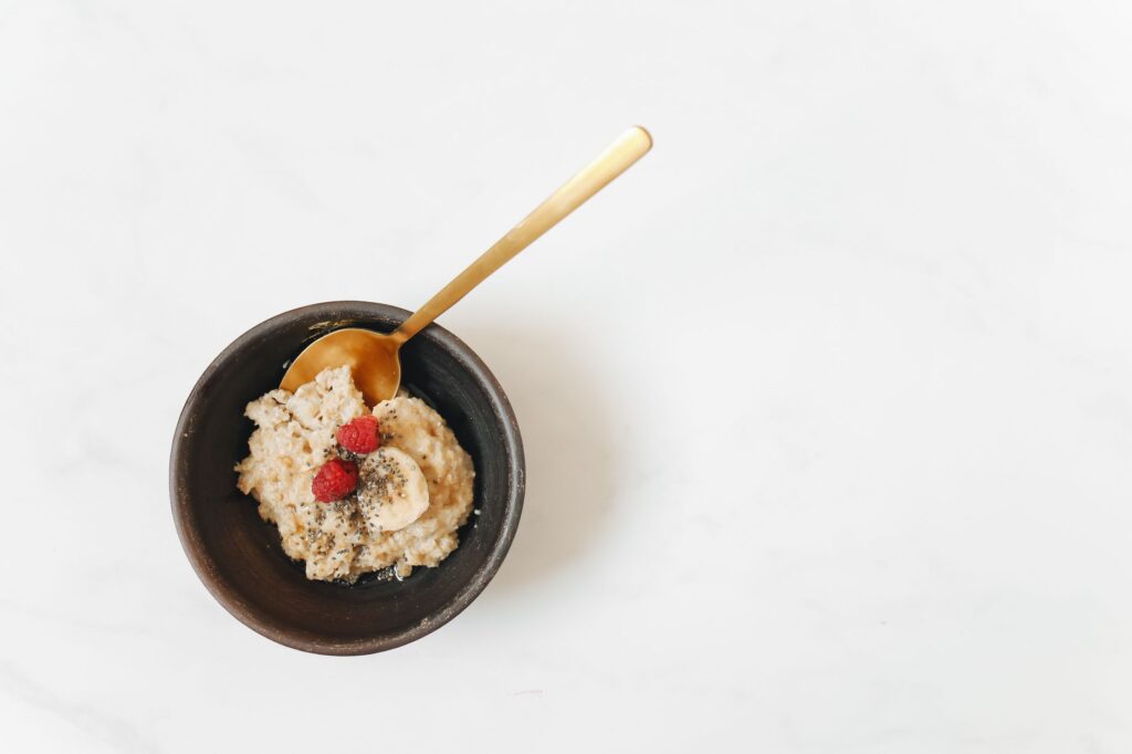 Vue aérienne d'un bol de petit-déjeuner avec des flocons d'avoine, des bananes, des framboises et des graines de chia sur une surface blanche.