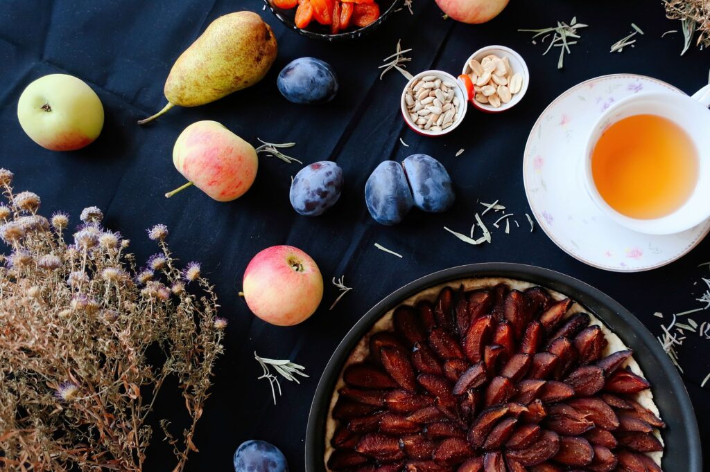 Composition de fruits colorés, tarte aux prunes et thé aux herbes sur une table sombre.