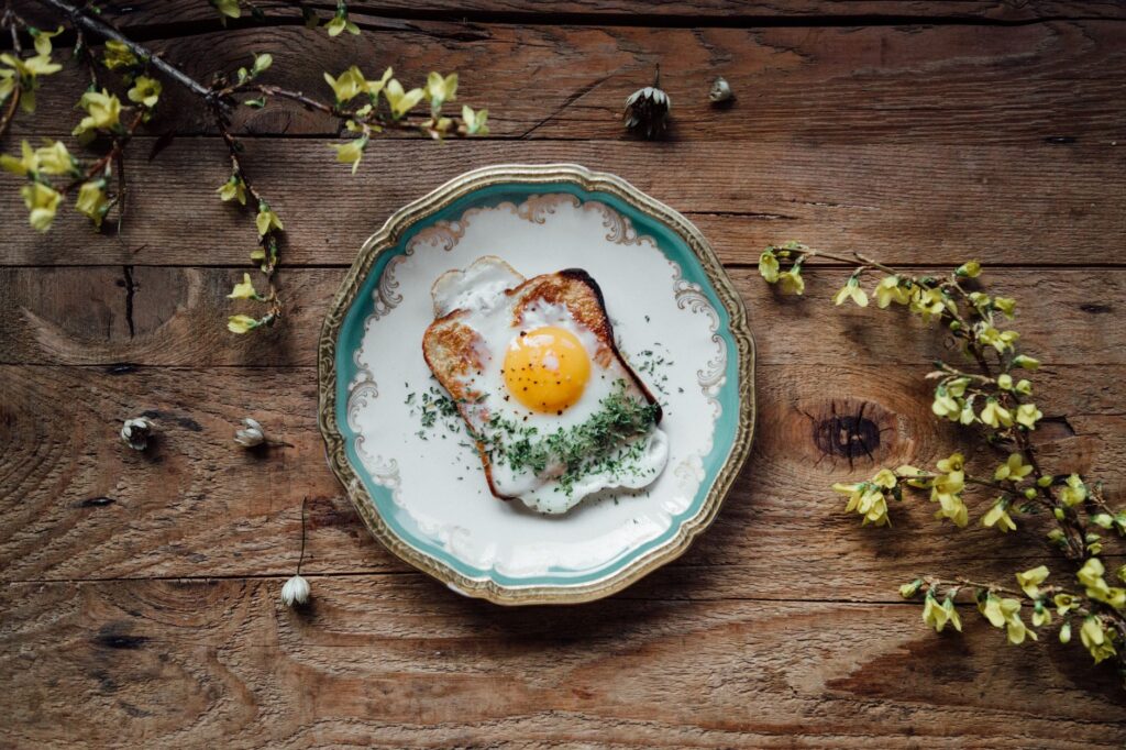 Œuf frit joliment présenté sur du pain grillé avec des herbes sur une table en bois rustique, entouré de fleurs jaunes.