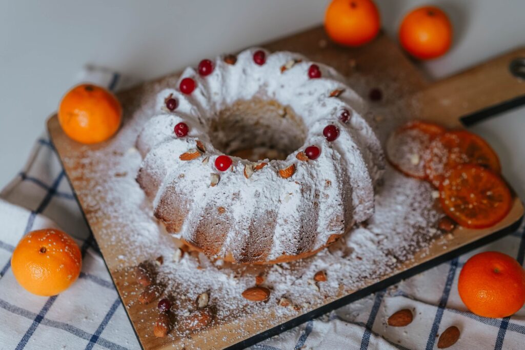 Un gâteau Bundt décoré de sucre glace, d'amandes et de fruits agrumes sur un plateau en bois.