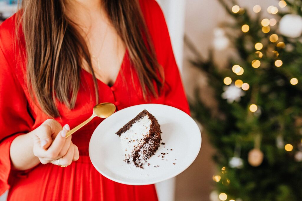 Une femme en robe rouge tient une part de gâteau au chocolat avec un sapin de Noël en arrière-plan.