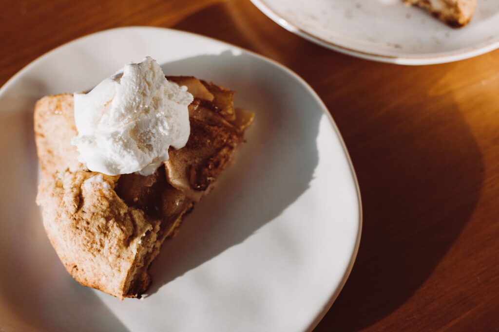 Gros plan sur une part de tarte aux pommes maison surmontée de crème fouettée dans une assiette en céramique.