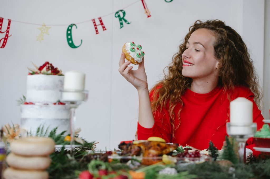 Femme en pull rouge savourant un dessert de Noël festif à l'intérieur, entourée de décorations de fête et de bougies.