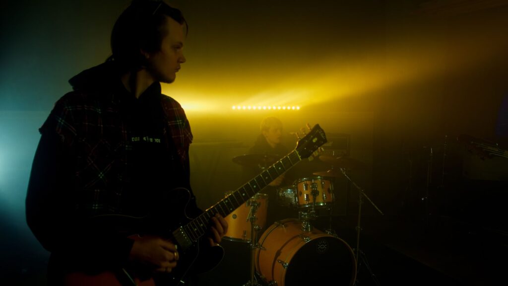 Musiciens se produisant avec des guitares et des tambours sous un éclairage scénique dramatique.