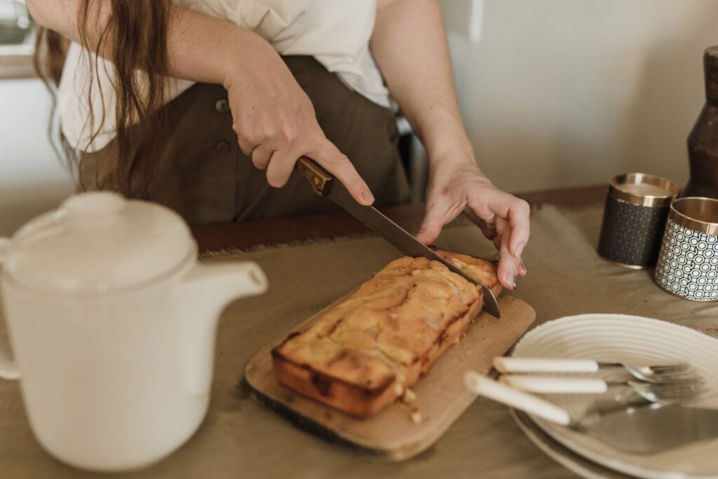 Femme tranchant un gâteau fraîchement cuit dans une atmosphère chaleureuse de cuisine avec une théière sur la table.