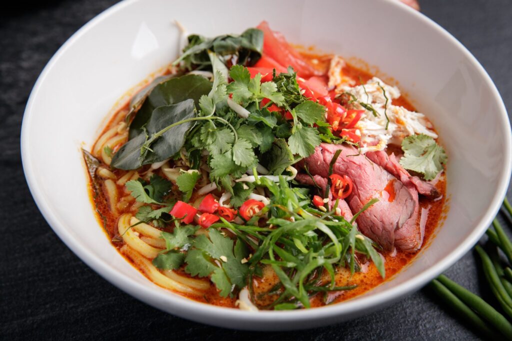 Une soupe de nouilles asiatique vibrante et épicée avec des herbes fraîches, du bœuf et des piments dans un bol blanc.