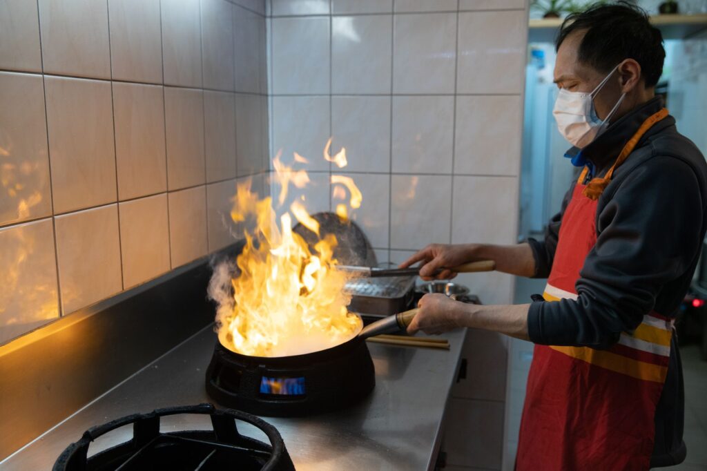 Un chef asiatique portant un masque facial prépare un plat flamboyant dans une cuisine commerciale.