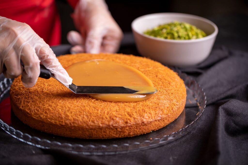 Un pâtissier utilise une spatule pour appliquer du caramel sur un dessert persan avec un bol de pistaches à proximité.
