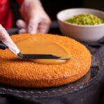 Un pâtissier utilise une spatule pour appliquer du caramel sur un dessert persan avec un bol de pistaches à proximité.