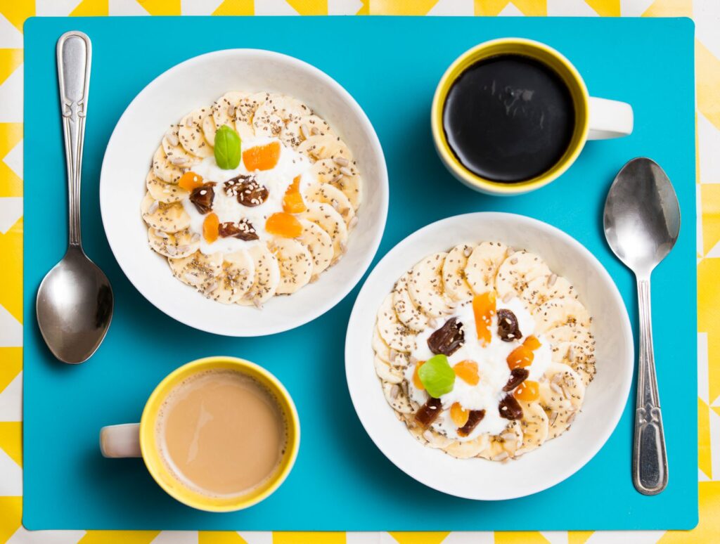 Vue de dessus de deux bols de petit déjeuner avec des bananes, du yaourt et des tasses à café sur un fond vibrant.