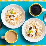 Vue de dessus de deux bols de petit déjeuner avec des bananes, du yaourt et des tasses à café sur un fond vibrant.
