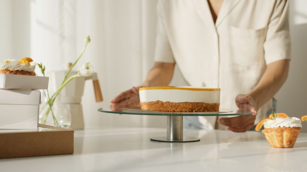 Un pâtissier arrange un gâteau fraîchement préparé sur un support en verre, avec des cupcakes à proximité, dans une cuisine lumineuse.