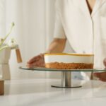 Un pâtissier arrange un gâteau fraîchement préparé sur un support en verre, avec des cupcakes à proximité, dans une cuisine lumineuse.