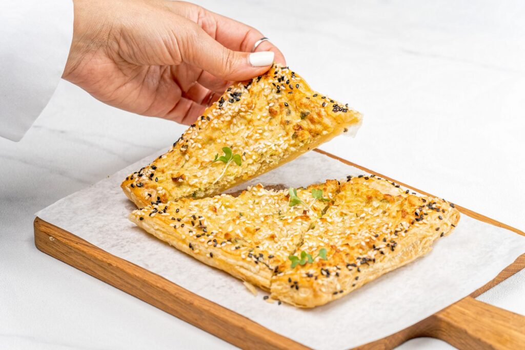 Gros plan sur une main qui prend une viennoiserie aux graines de sésame sur une planche en bois, mettant en valeur une délicieuse photographie culinaire.