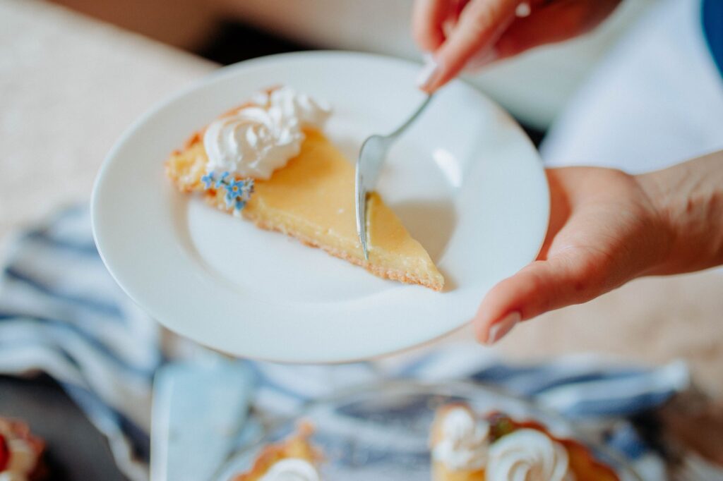 Gros plan d'une main tenant une part de tarte au citron garnie de crème et de fleurs sur une assiette.