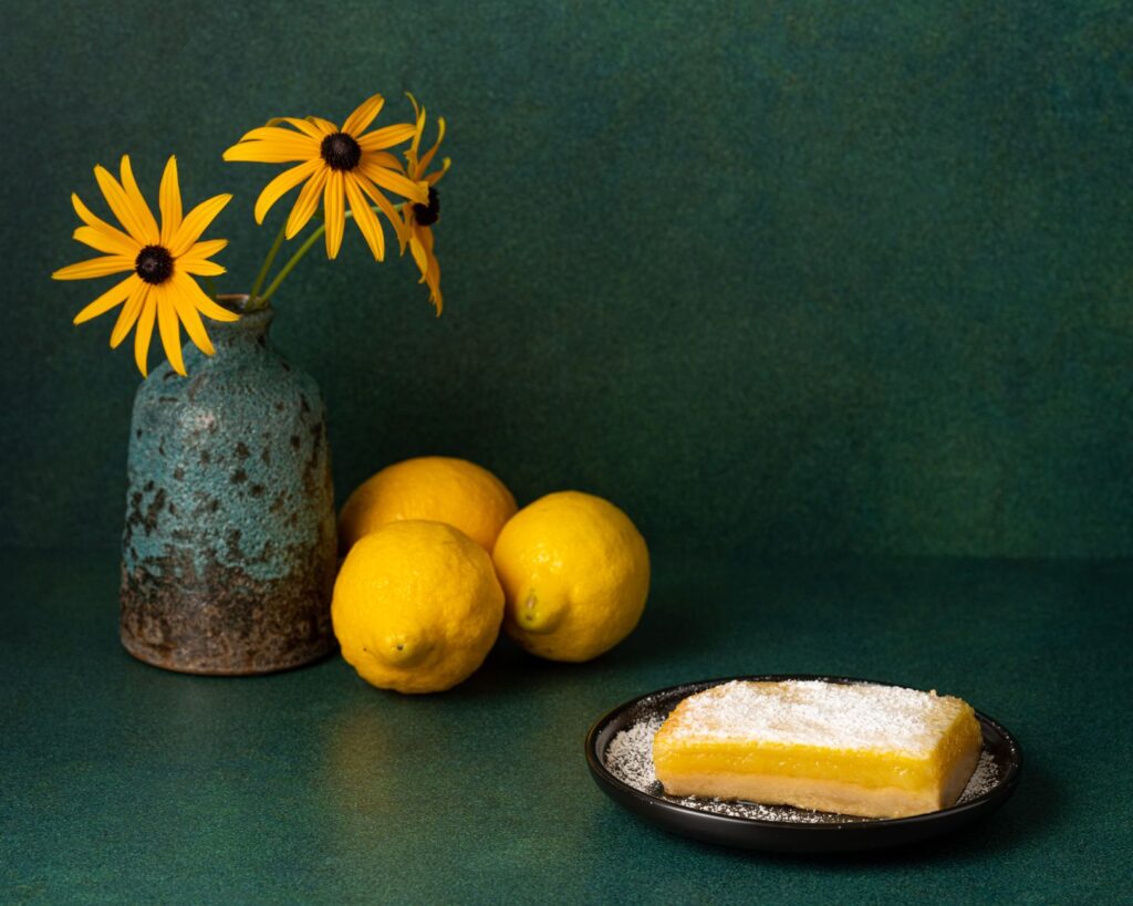 Une tarte au citron saupoudrée de sucre à côté d'un vase rustique avec des fleurs jaunes sur un fond vert.