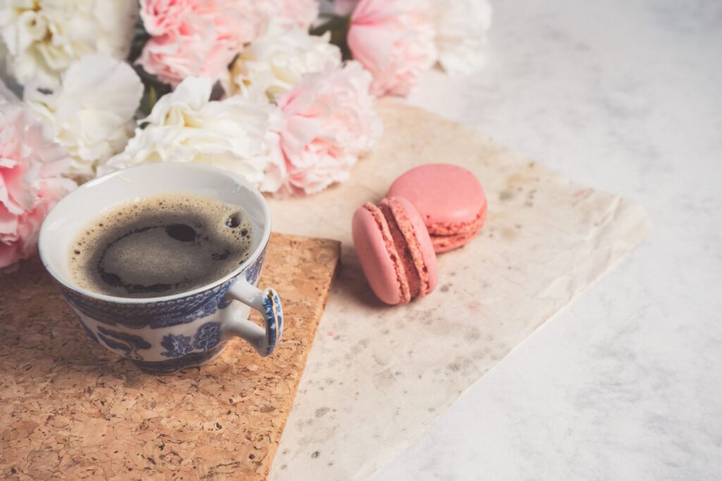 Une mise en scène charmante avec une tasse de café, des macarons roses et des fleurs pastel douces.