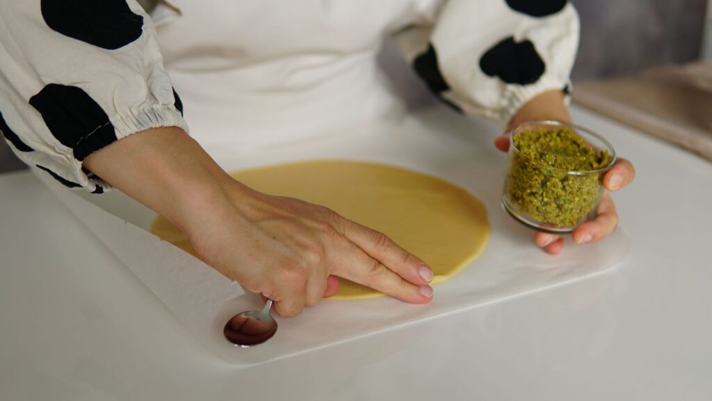 Mains préparant de la pâte avec une garniture de pistache sur du papier sulfurisé.