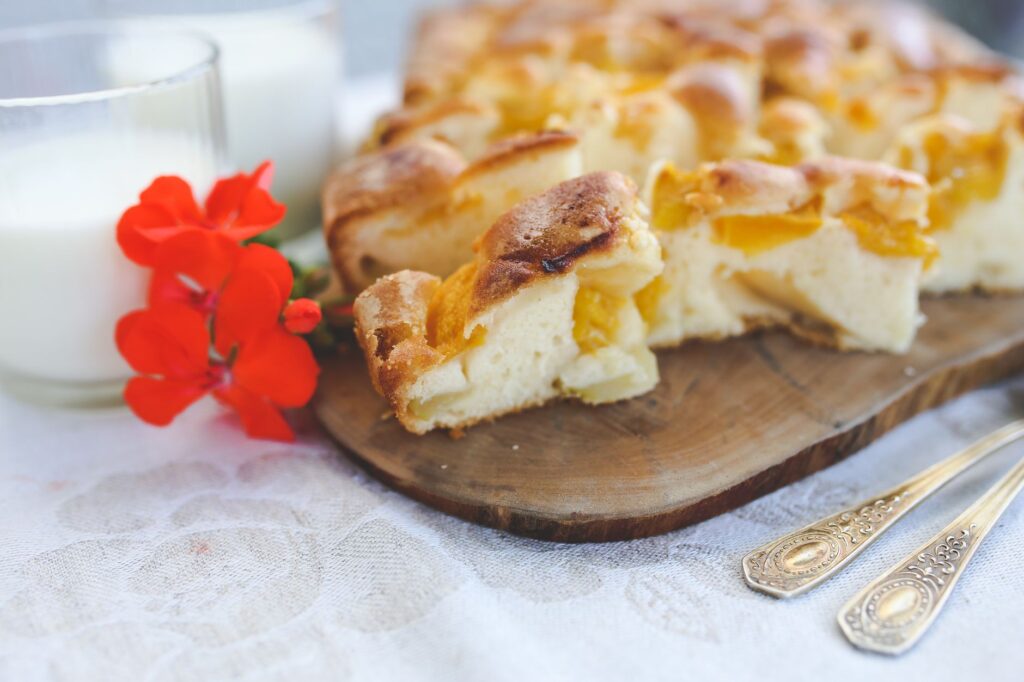 Une part de tarte aux pêches maison sur une planche en bois avec du lait et des fleurs rouges.