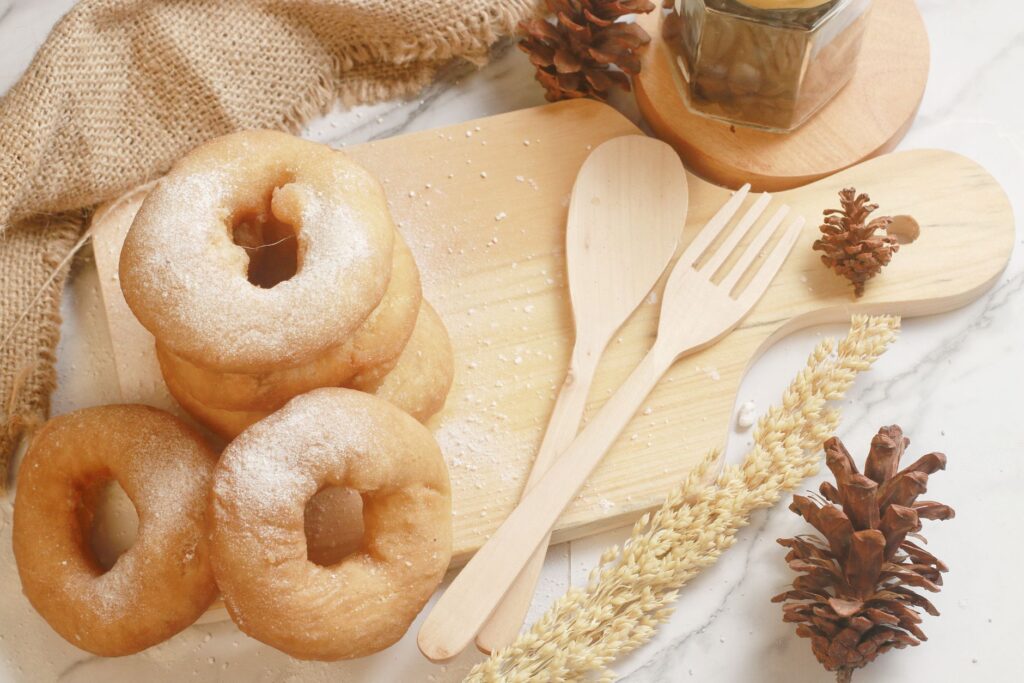 Une pile de beignets poudrés sur une planche en bois rustique avec des pommes de pin et des ustensiles de cuisine pour une ambiance chaleureuse.