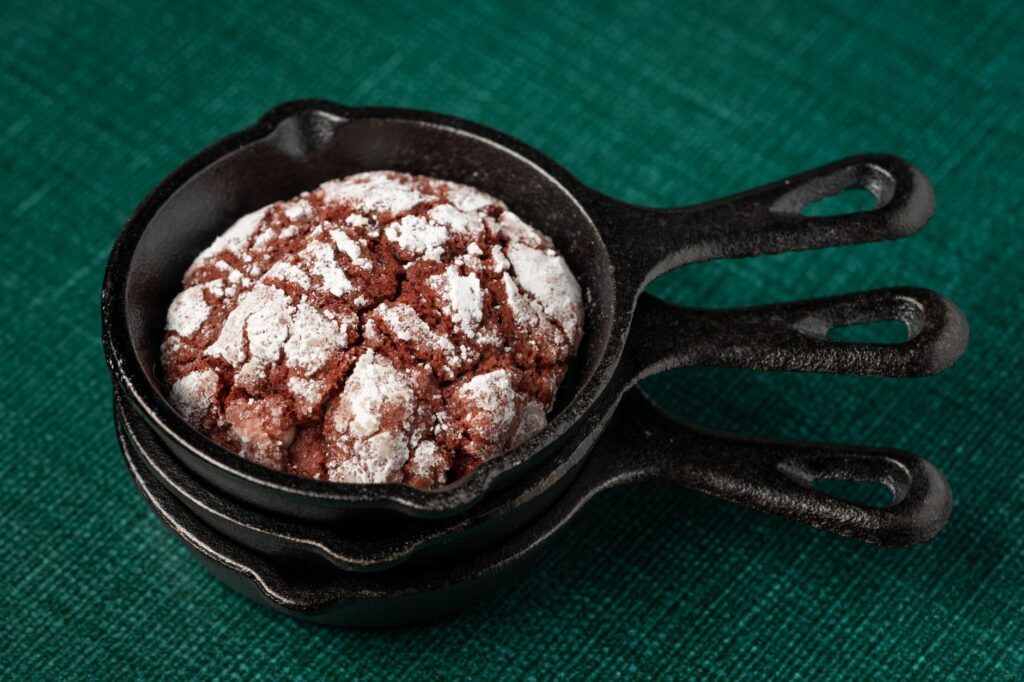 Gros plan sur un cookie au chocolat fripé dans une petite poêle noire sur un fond vert texturé.