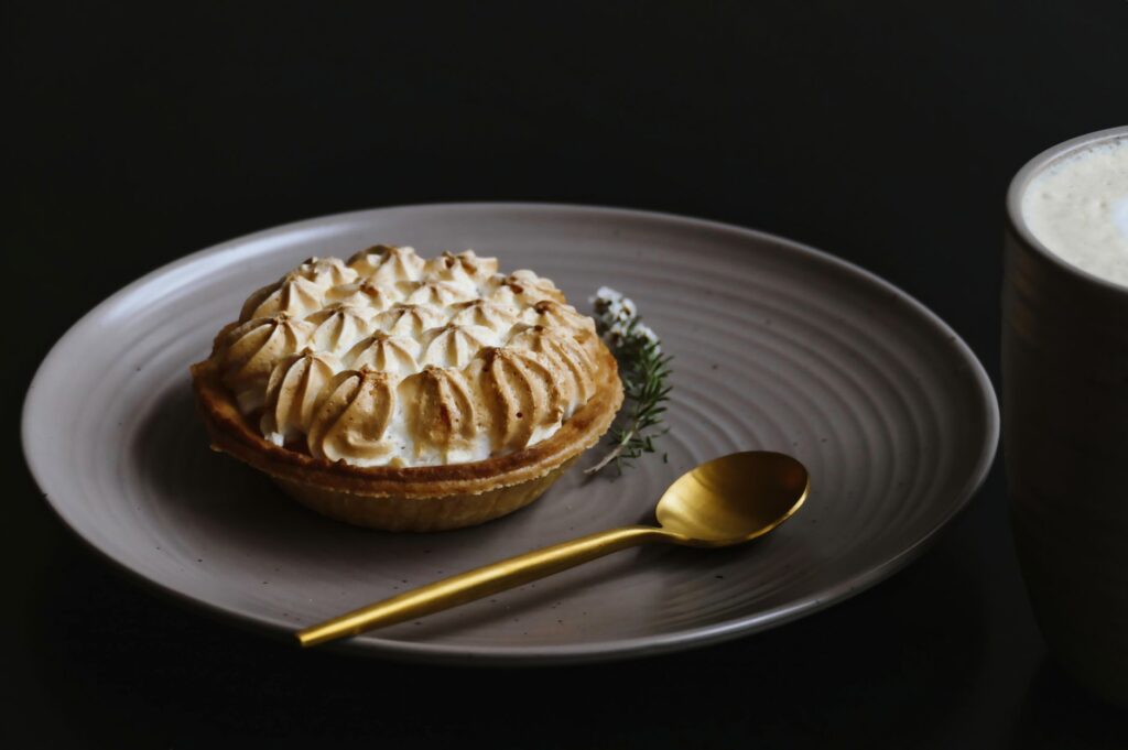 Une tarte meringuée au citron magnifiquement présentée sur une assiette avec une cuillère dorée et une tasse, parfaite pour un petit-déjeuner gourmet.