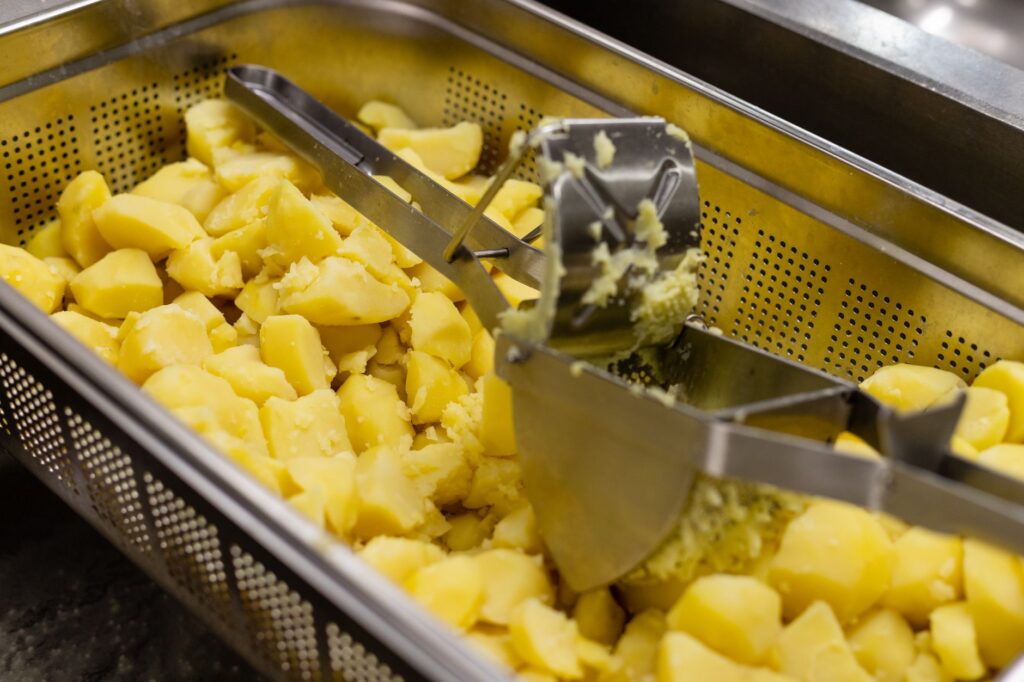 Des pommes de terre jaunes fumantes écrasées avec un presse-purée en métal dans un cadre de cuisine professionnelle.
