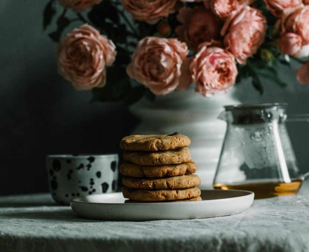 Des cookies aux pépites de chocolat empilés sur une assiette avec des fleurs roses et du thé. Nature morte chaleureuse.