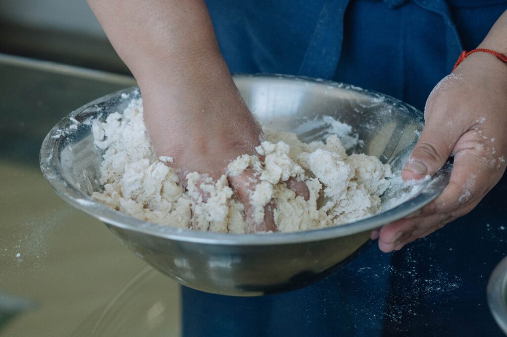 Gros plan sur des mains pétrissant de la pâte dans un bol en acier inoxydable, illustrant le processus de cuisson.