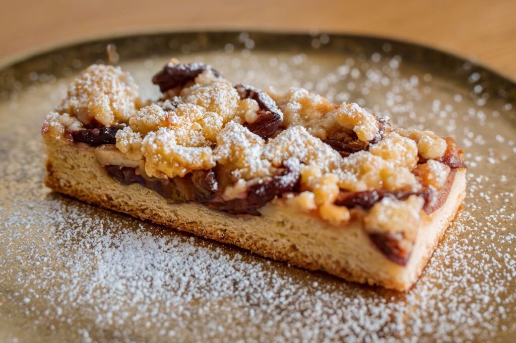 Une tranche de gâteau crumble à la prune sur une assiette saupoudrée de sucre glace.