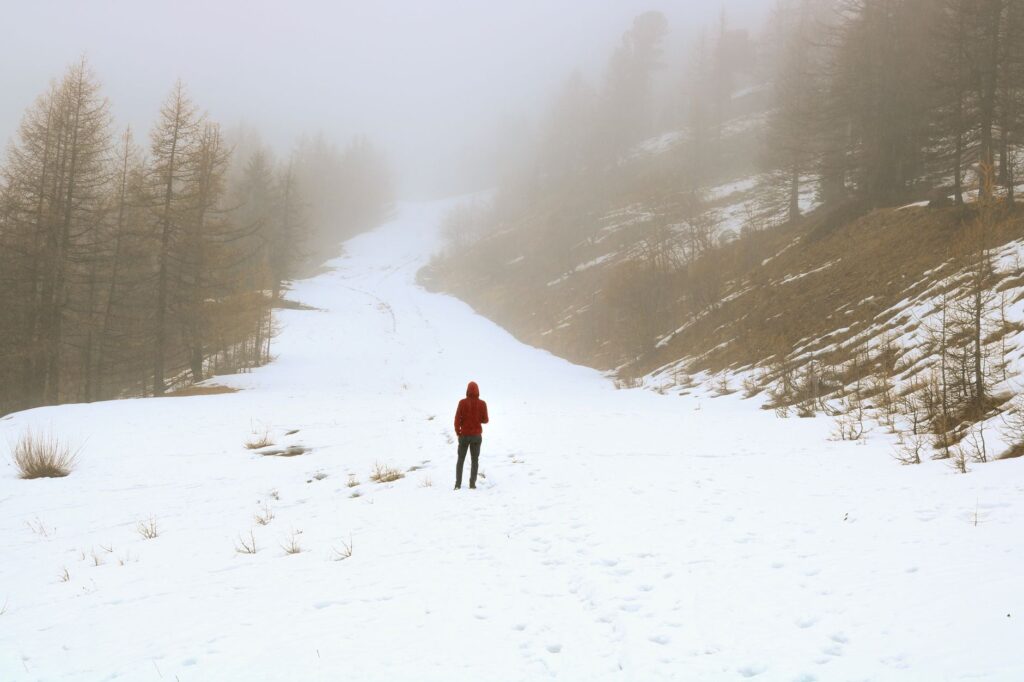 Une personne seule, vêtue de rouge, explore un sentier de montagne brumeux et enneigé, entouré d'arbres.