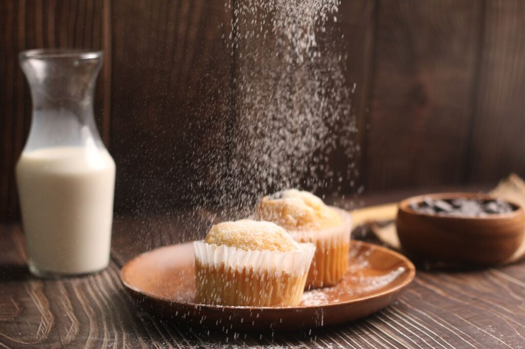 Muffins fraîchement cuits saupoudrés de sucre, accompagnés de lait, dans une ambiance de cuisine rustique.