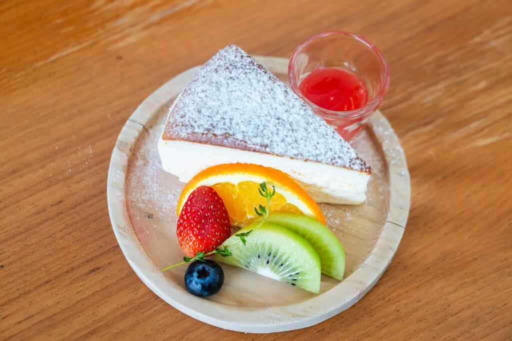 Une part de gâteau au fromage saupoudrée de sucre glace et accompagnée de fruits frais, servie sur une assiette en bois.