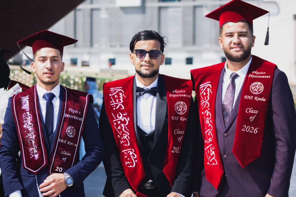 Trois jeunes diplômés de l'ENS Kouba d'Alger, coiffés de leurs toques et vêtus de leurs robes de cérémonie, posent avec enthousiasme pour une photo.