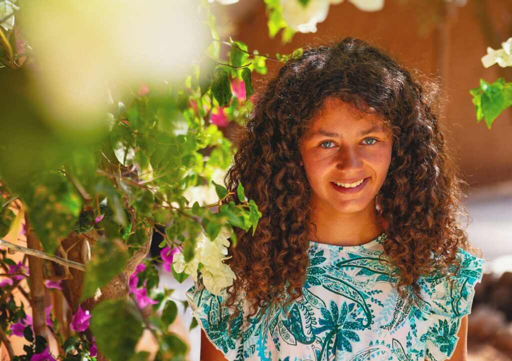 Une jeune fille souriante entourée d'un feuillage coloré dans un jardin ensoleillé.