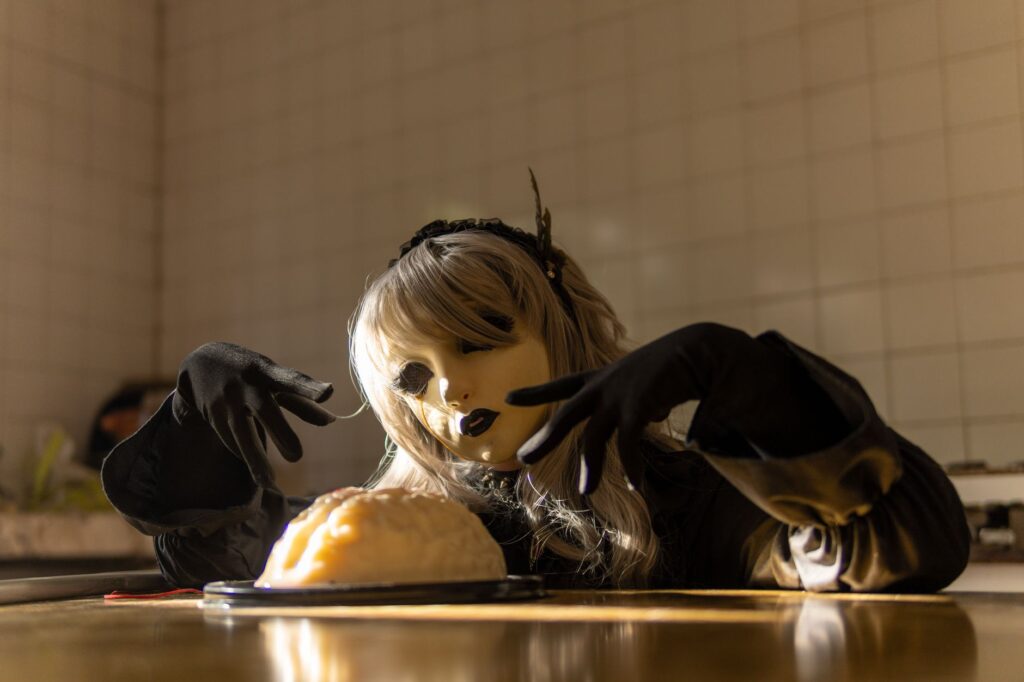Une personne portant un masque et un costume de poupée pose de façon étrange avec un gâteau en forme de cerveau, évoquant le thème d'Halloween.