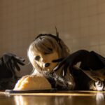 Une personne portant un masque et un costume de poupée pose de façon étrange avec un gâteau en forme de cerveau, évoquant le thème d'Halloween.