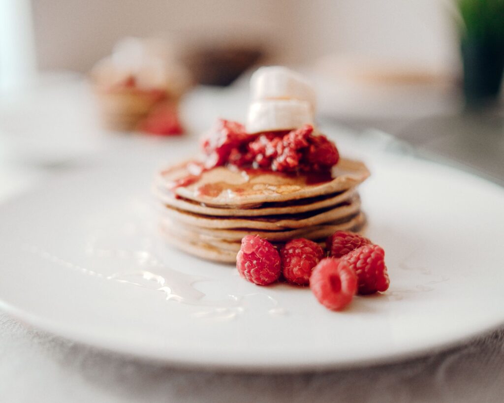 Pile de crêpes garnies de framboises fraîches et de sirop sur une assiette blanche.