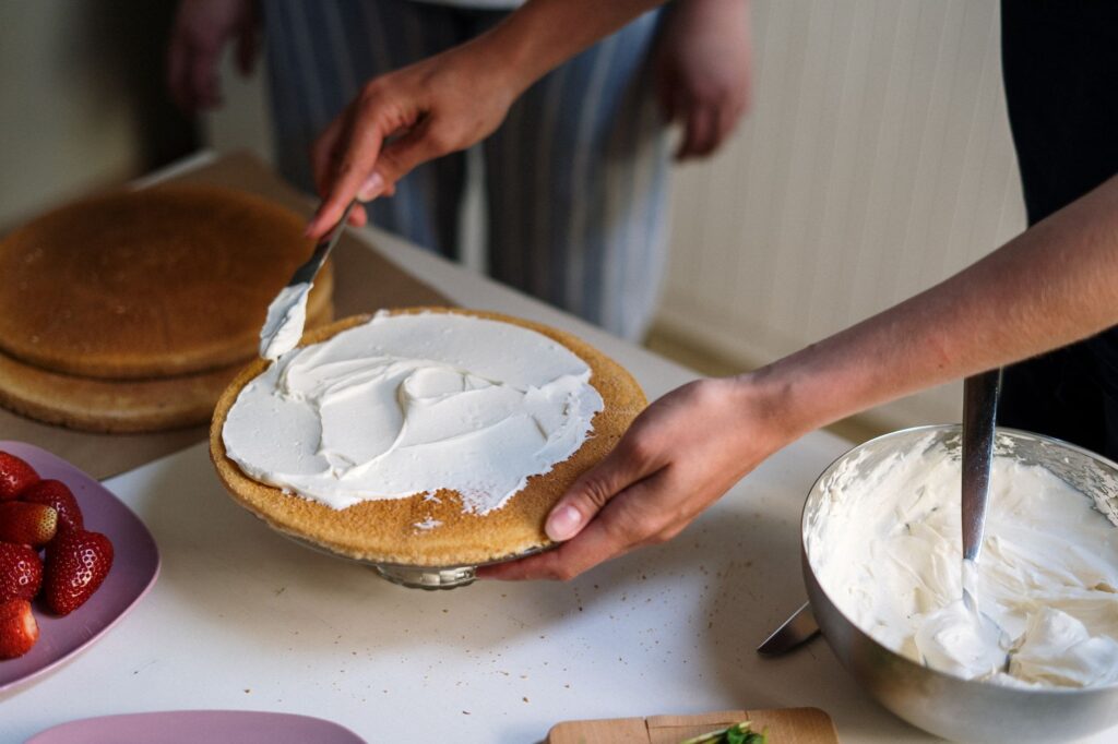 Mains préparant un gâteau avec un glaçage à la crème dans une cuisine confortable, featuring des fraises et un saladier.