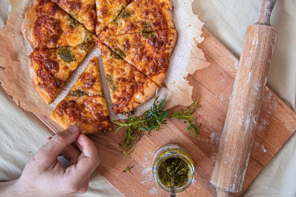 Main tendue vers une part de pizza maison fraîchement cuite avec des herbes sur une planche en bois.